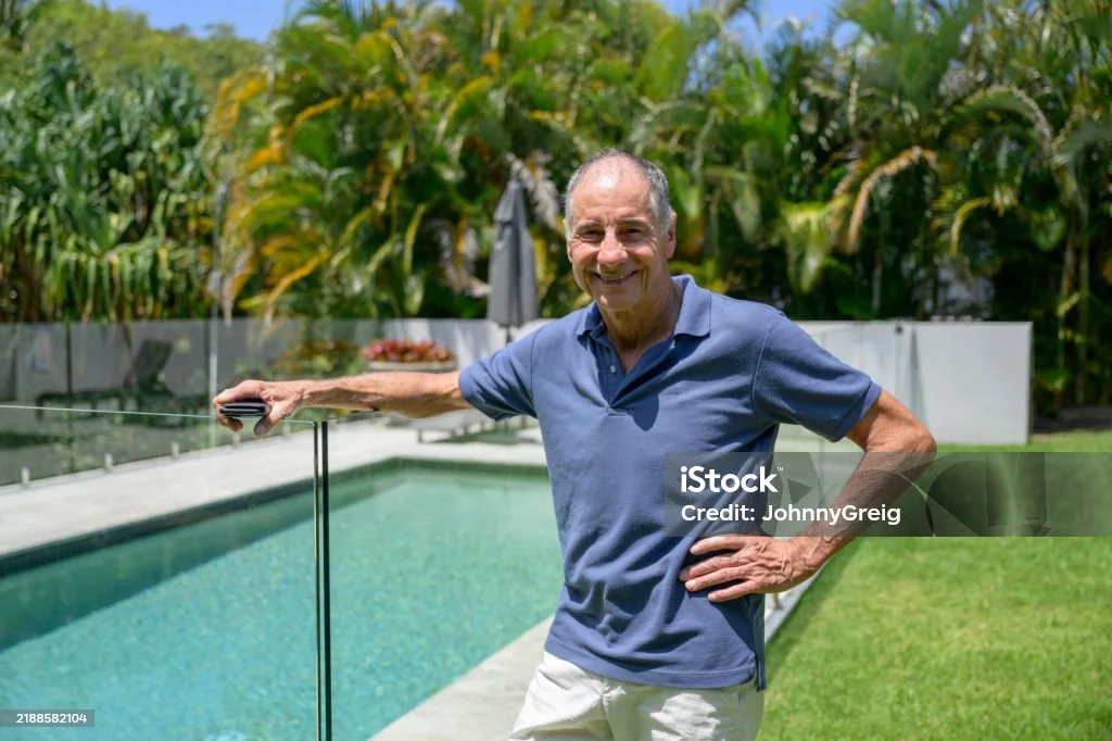 A smiling retired man holding his phone beside his fenced backyard pool